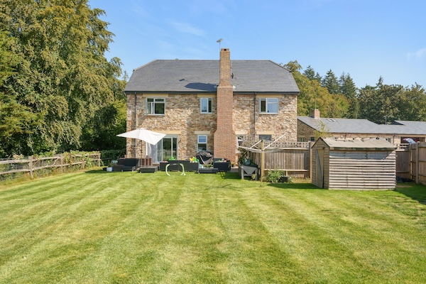 A detached stone house with a large garden, patio seating, a wooden shed, and a rotary clothesline, set against trees and neighbouring houses.