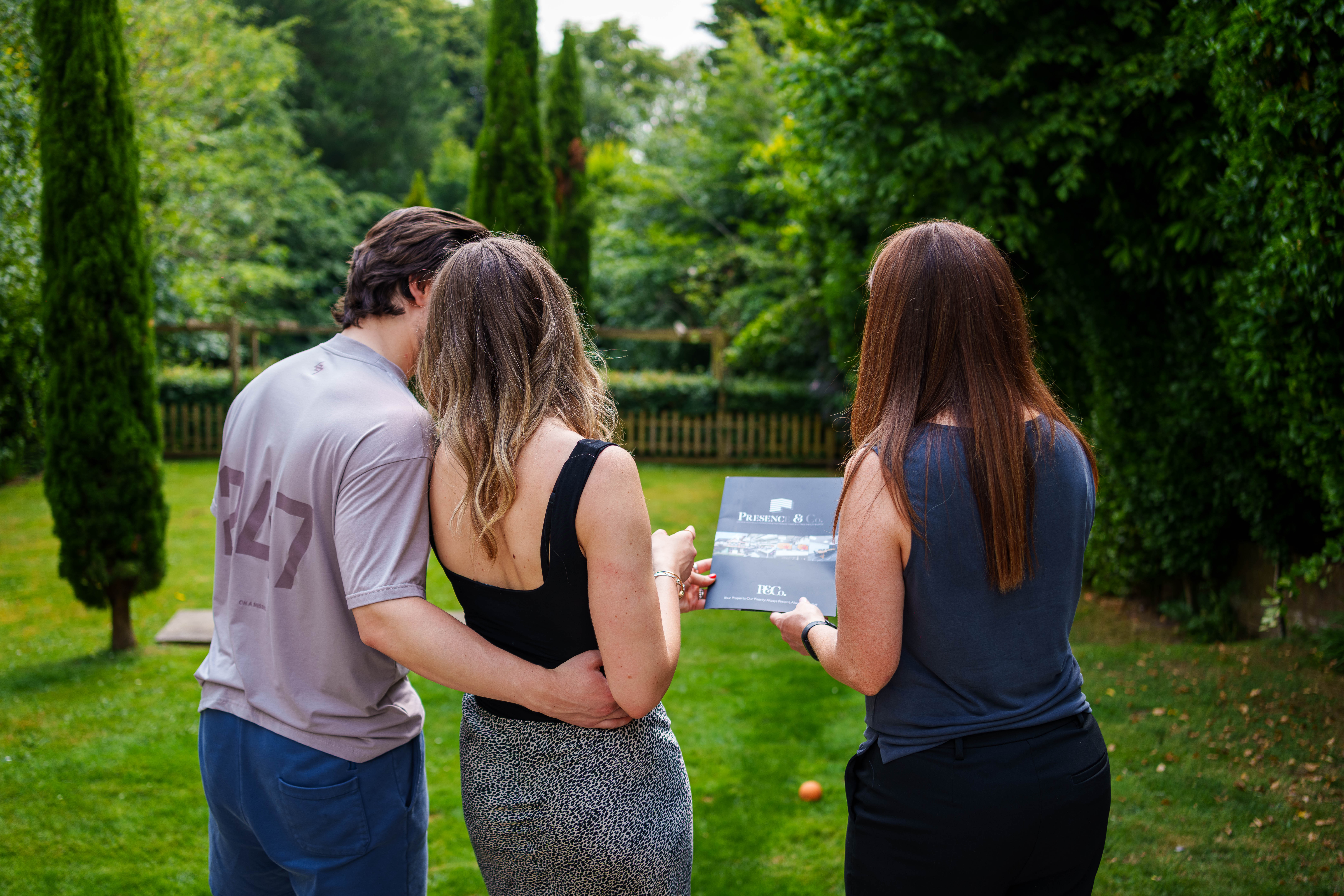 A couple having a viewing in the garden on a summers day with his arm around her waist and estate agent showing Presence & Co book