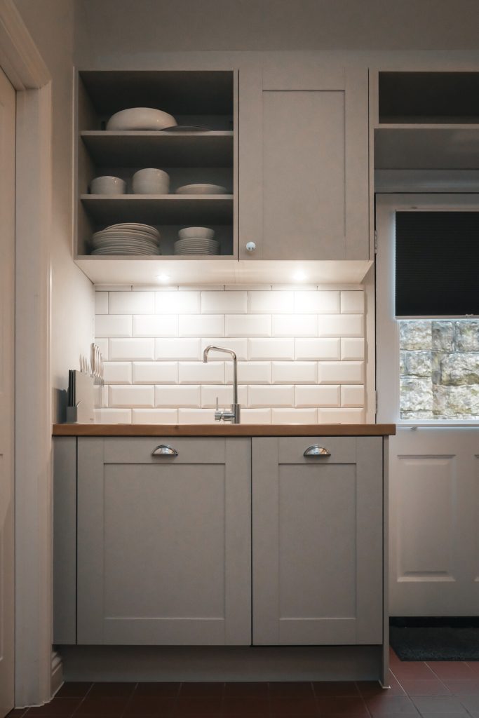 kitchen interior with grey wooden cuploards, warm low lit lighting underneath the top cupboards, plates and bowls places on the shelves. Bright white tiles to compliment the colour of the cupboard doors.