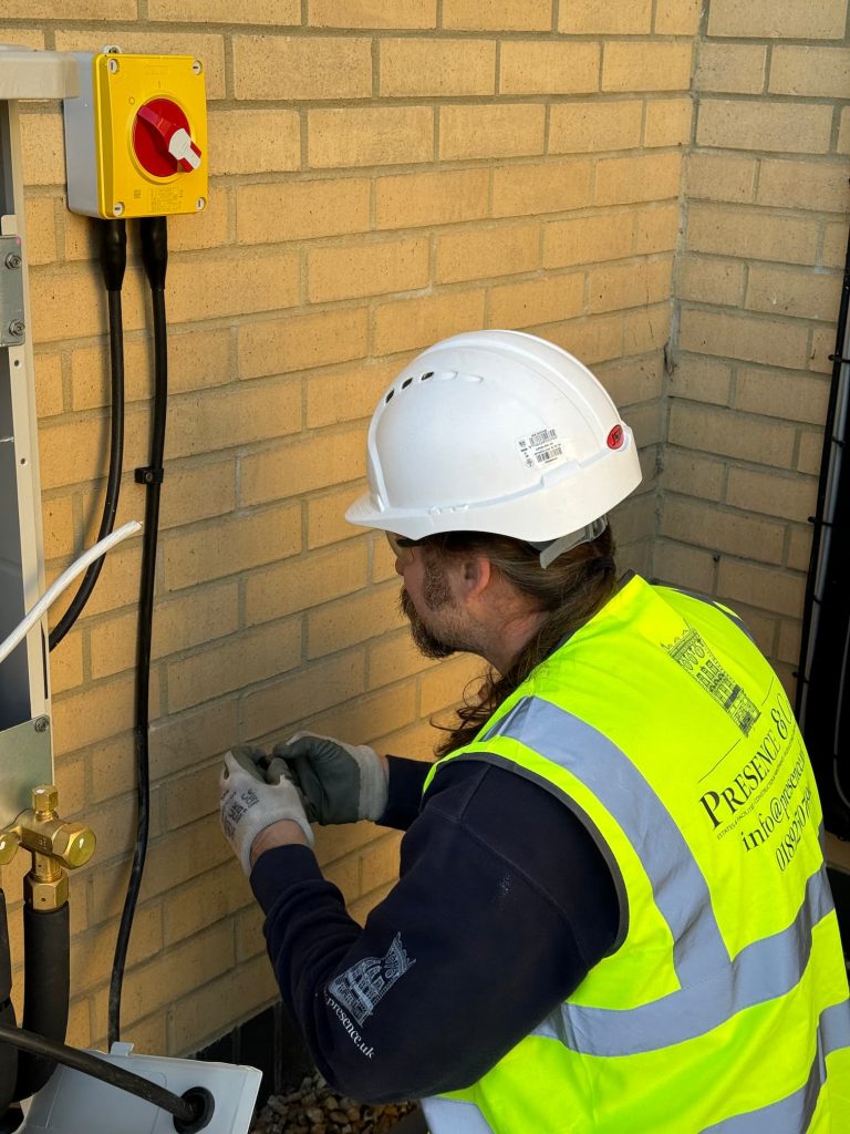 Electrician knelt on the floor outside, wearing a presence & co. high vis and a white hard hat for PPE. Working on re wiring an external fixture 