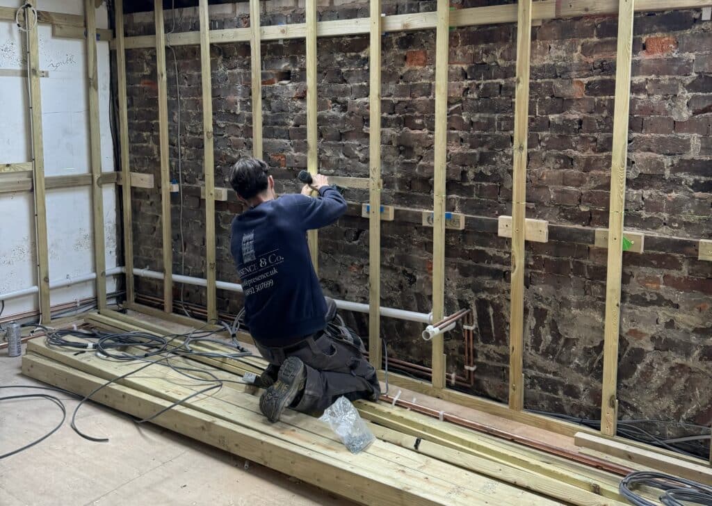 Electrician knelt on the ground wearing a navy blue Presence & Co. jumper, holding a power tool and drilling into the batten installation.