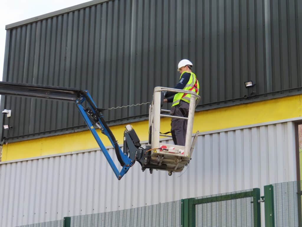electrician on a blue cherry picker outside a black and grey building, wearing a hard hat, green high vis and red harness. electrician using controller to go up and down