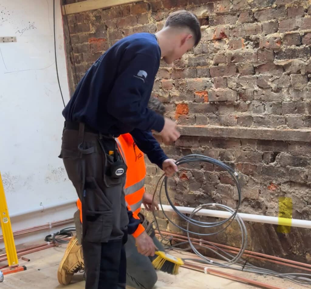 Electrician standing on site wearing a navy blue Presence & Co. jumper, standing next to another electrician wearing an orange highvis. Electrician coiling cables for first fix.