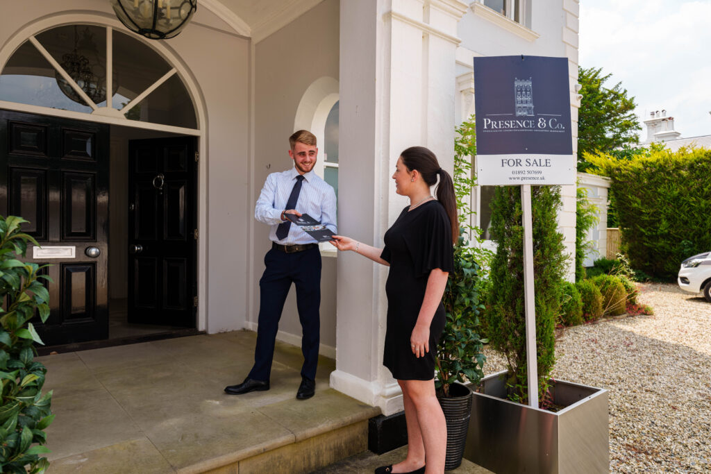 estate agent stood outside property wearing a suit and blue tie, handing a brochure to a woman with a black dress on ready to look around the property. stood in a large archway at the front of the building. presence &co. for sale sign in the back
