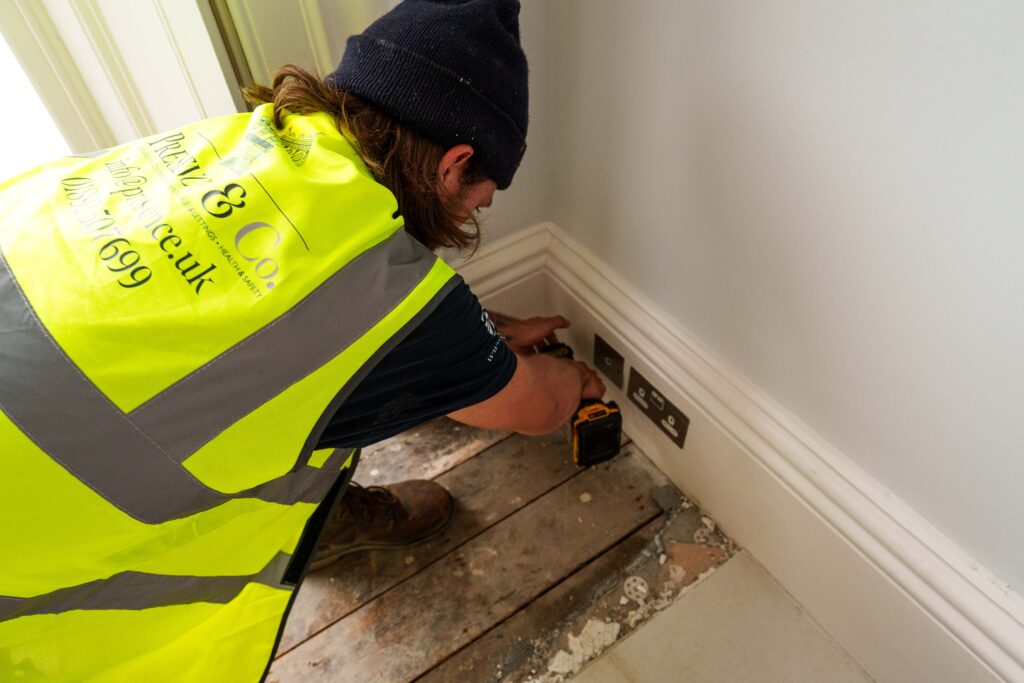 Carpenter wearing yellow hivis jacket and kneeling on the floor to fit wooden flooring. Carpenter is wearing Presence & Co. jacket, a blue beanie and holding a screwdriver. 