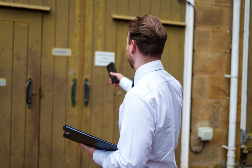man in a white shirt taking pictures of a wooden building door, holding a black notepad  and mobile phone 
