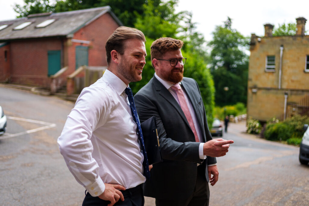 two men assesing a building, one wearing a white shirt and one wearing a blue jacket with a pink tie. standing outside the building with trees in the background 