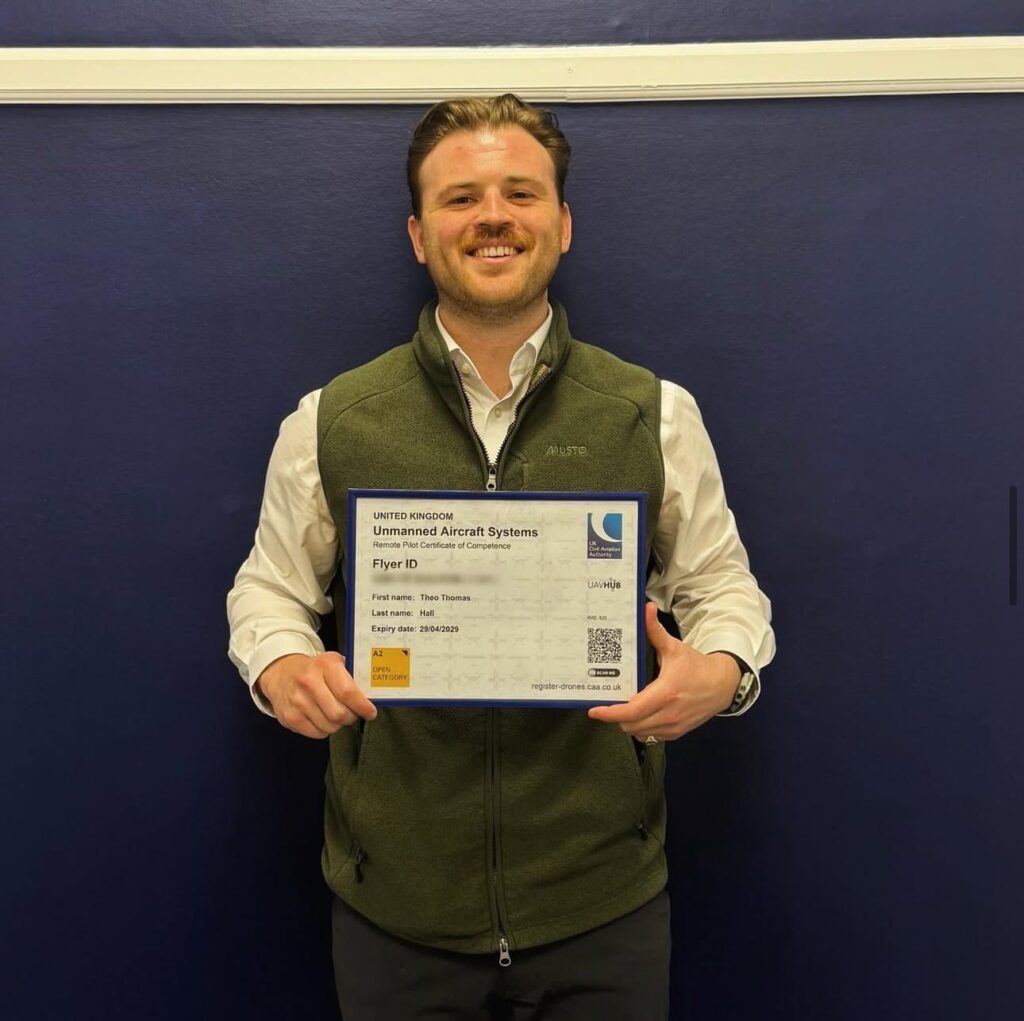 Theo hall standing in front of a navy blue wall wearing a green jacket and holding a white certificate 