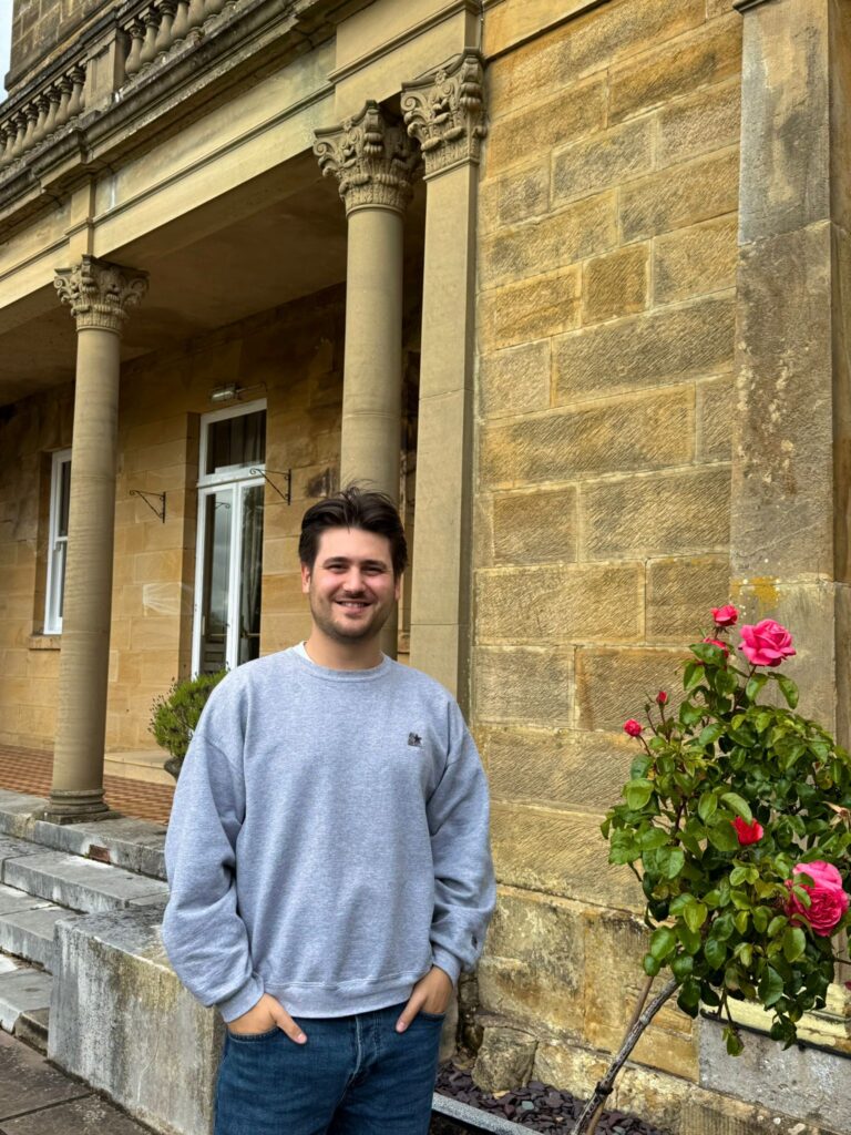 Sam, Administrator, stoof outside a yellow brick building with pink roses in the background