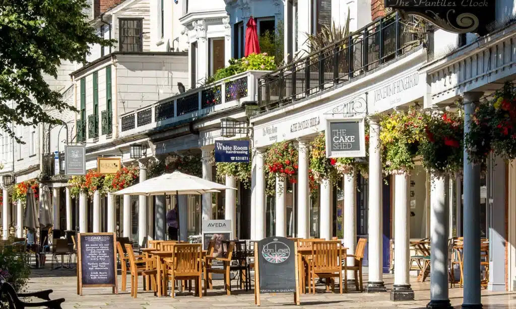 tunbridge wells pantiles in the sun, featuring shops and cafes, surrounded by trees and hanging flowers and tables and chairs outside. written menu boards 
