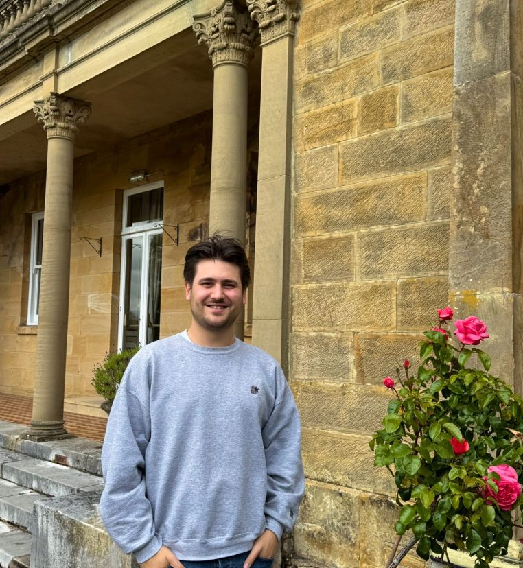 Sam, Administrator, stoof outside a yellow brick building with pink roses in the background